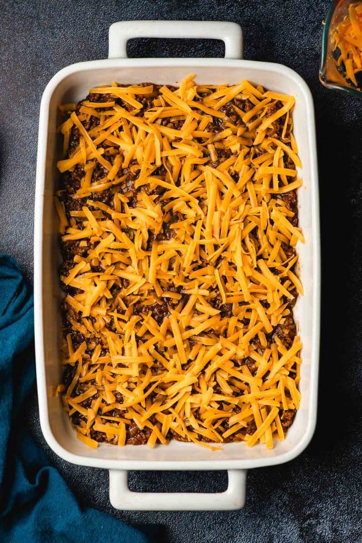 Shredded cheddar cheese on top of sloppy joe mix in a white casserole dish.
