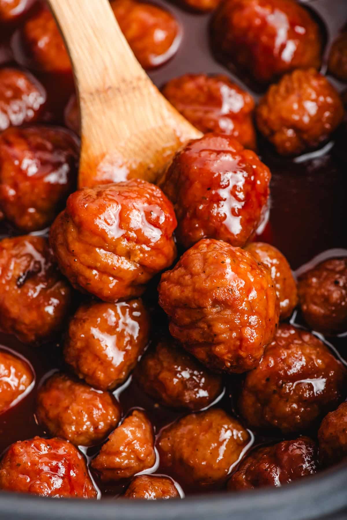 Wooden spoon scooping up grape jelly meatballs from a slow cooker.