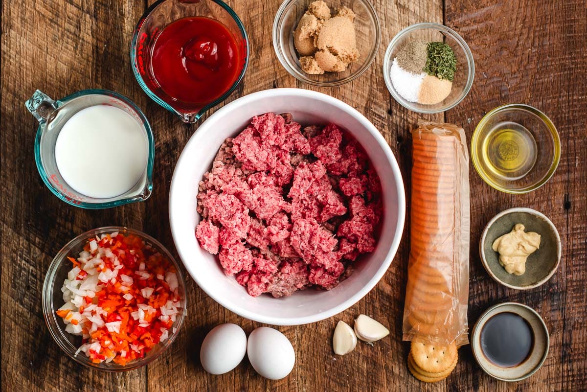 Ingredients for meatloaf on a wood background: ritz crackers, bell pepper, onion, eggs, garlic, milk, ketchup, brown sugar, worcestershire, dijon mustard, and seasonings.