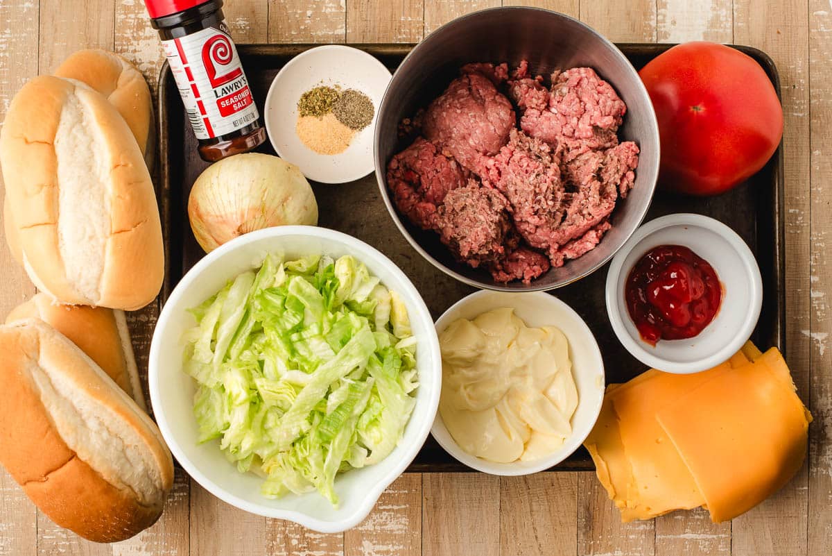 Ingredients for chop cheese on a cutting board: sub buns, Lawry's seasoning, onion, salt, pepper, garlic, ground beef, ketchup, mayonnaise, American cheese, and shredded lettuce.
