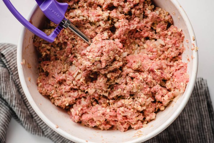 Uncooked meatball mix in a mixing bowl with a cookie scoop scooping one out.