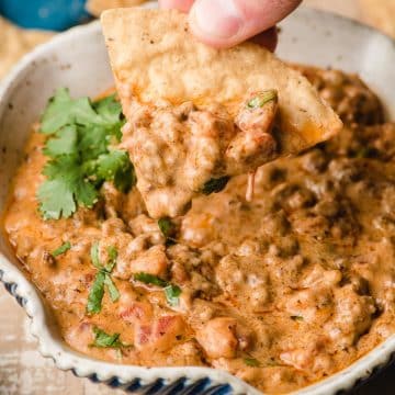 A chip scoops some ground beef queso dip out of a bowl.
