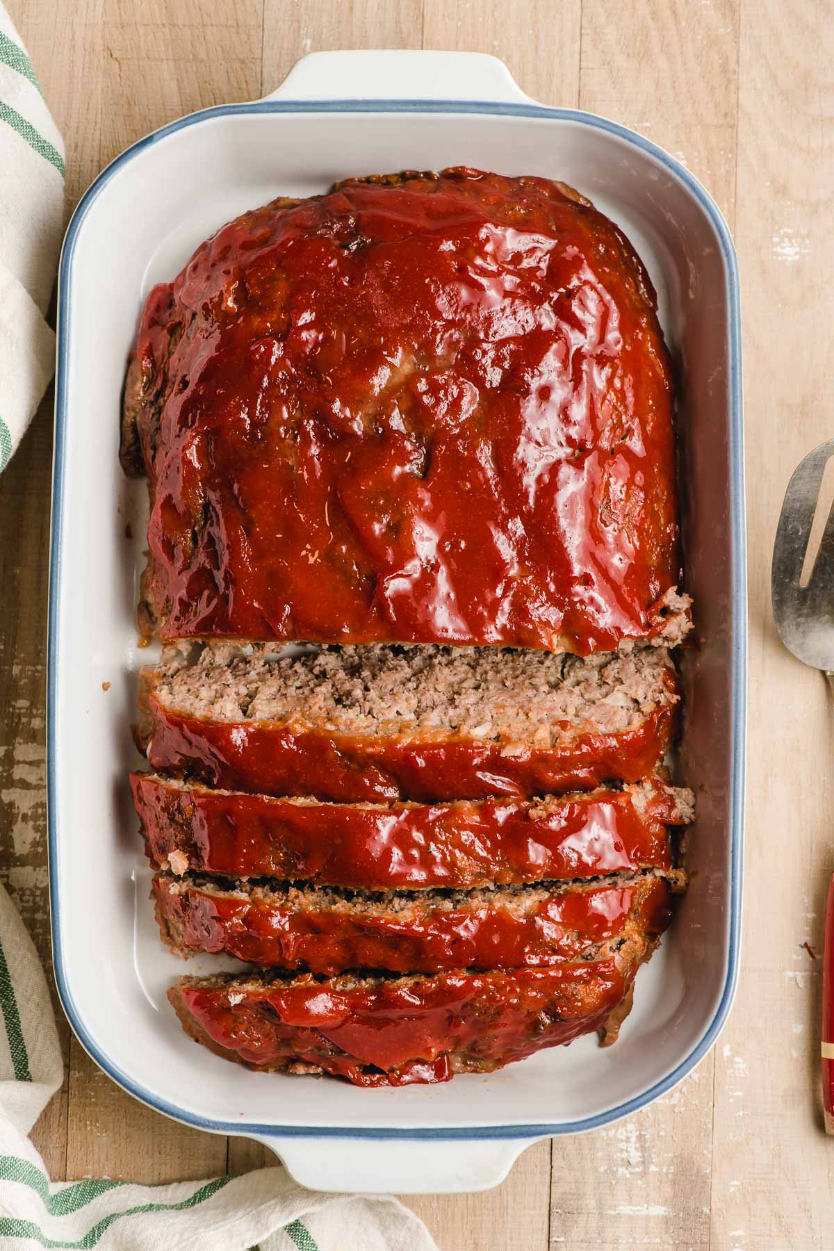 White casserole dish with a ketchup glazed meatloaf inside, half of it sliced and half whole.