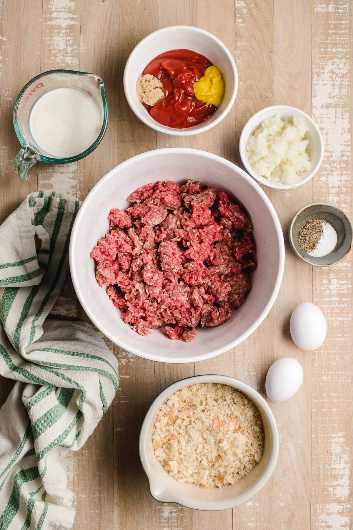Ingredients for meatloaf displayed in small bowls- ground beef, diced onions, eggs, breadcrumbs, milk, ketchup, and mustard.