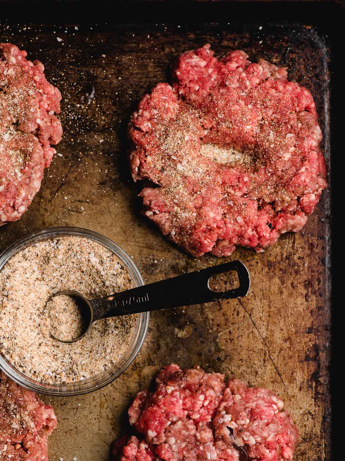Raw burger patties sprinkled with seasoning on a sheet pan.