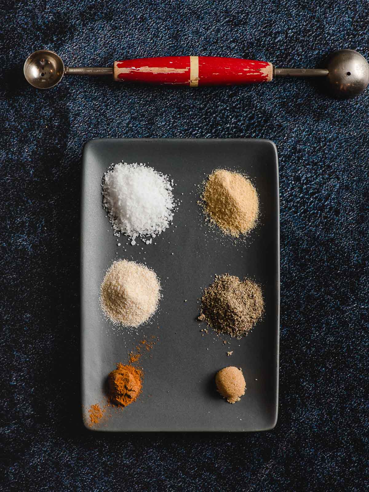 Spices for burgers shown in neat piles on a dark gray plate--salt, garlic powder, onion powder, pepper, cayenne pepper, brown sugar.