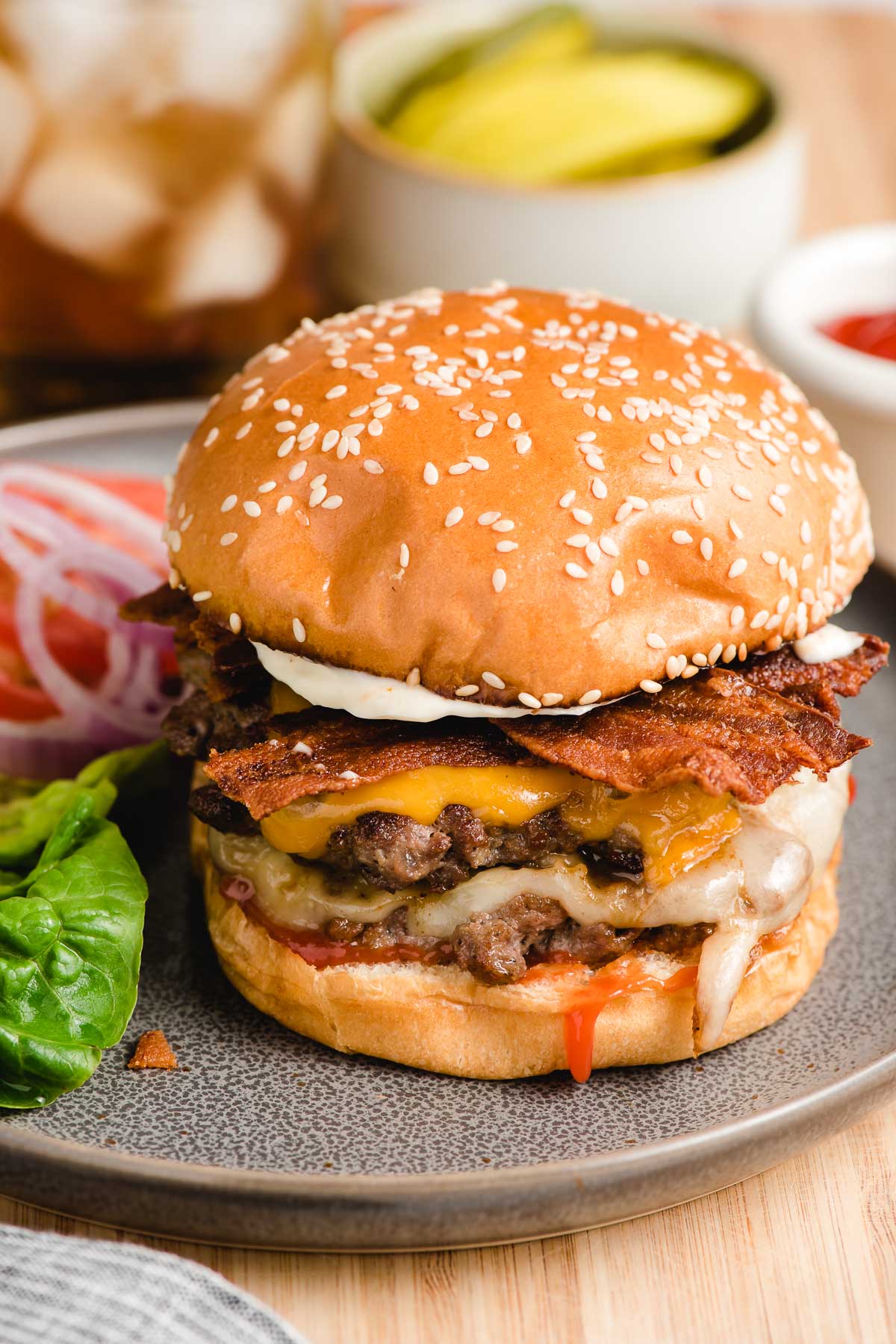 Double cheeseburger with bacon shown on a gray plate surrounded by toppings like lettuce, tomato, onion, and pickles.