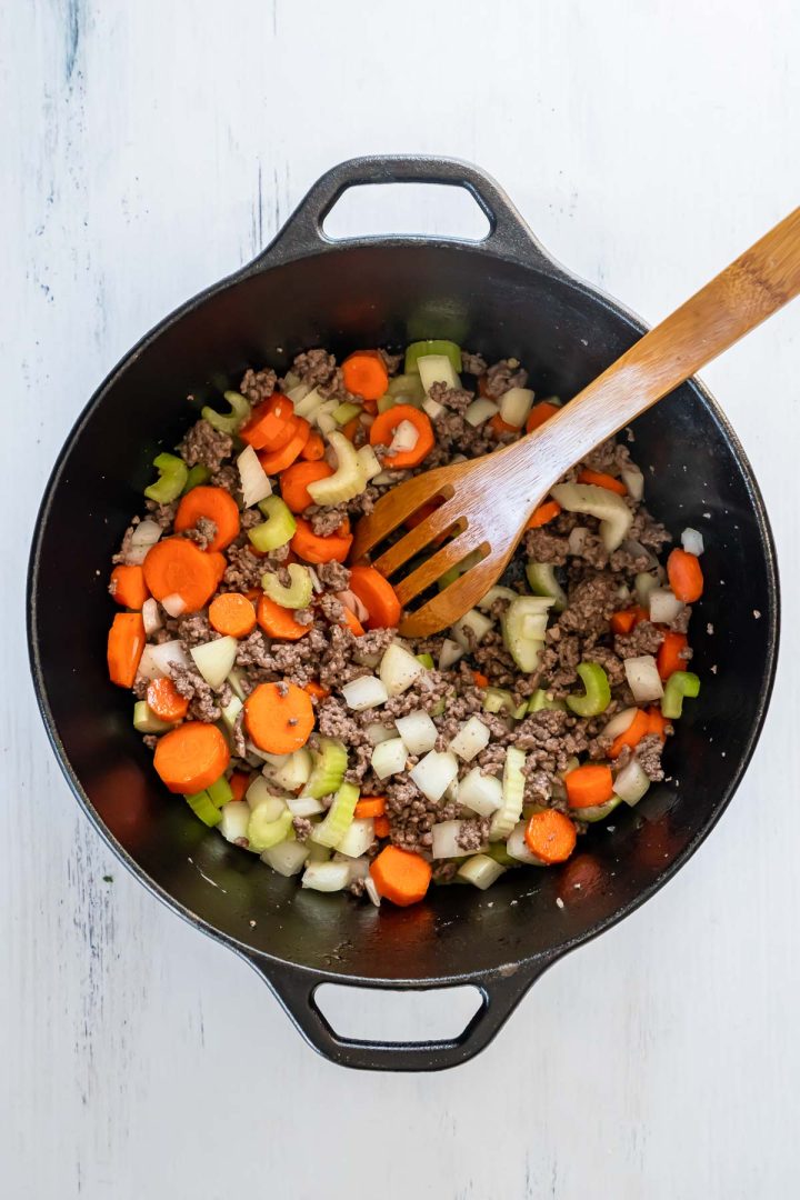 Wooden spoon stirring together ground beef, onions, carrots, and celery in a black dutch oven.