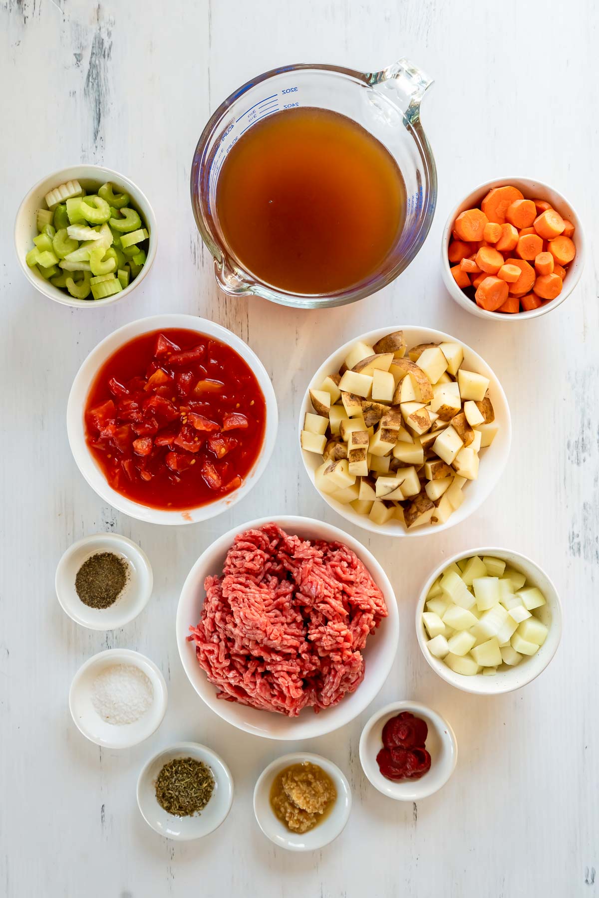 Ingredients for ground beef soup arranged in bowls--ground beef, chopped onions, potatoes, celery and carrots, beef broth, tomato paste, salt, pepper, diced tomatoes, and seasonings.,
