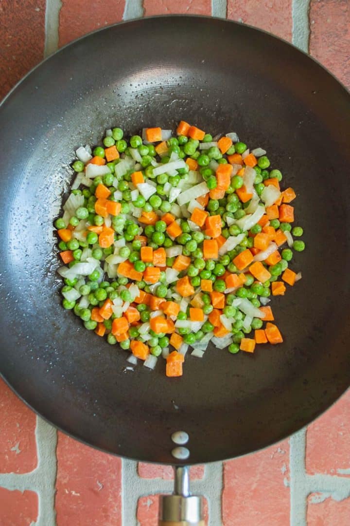 Chopped onion, peas, and carrots in a black saute pan.