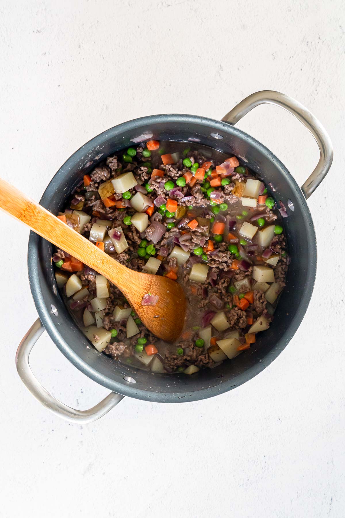 Pot full of cooked ground beef, diced potatoes, carrots, peas, and onions, being stirred by a wooden spoon.