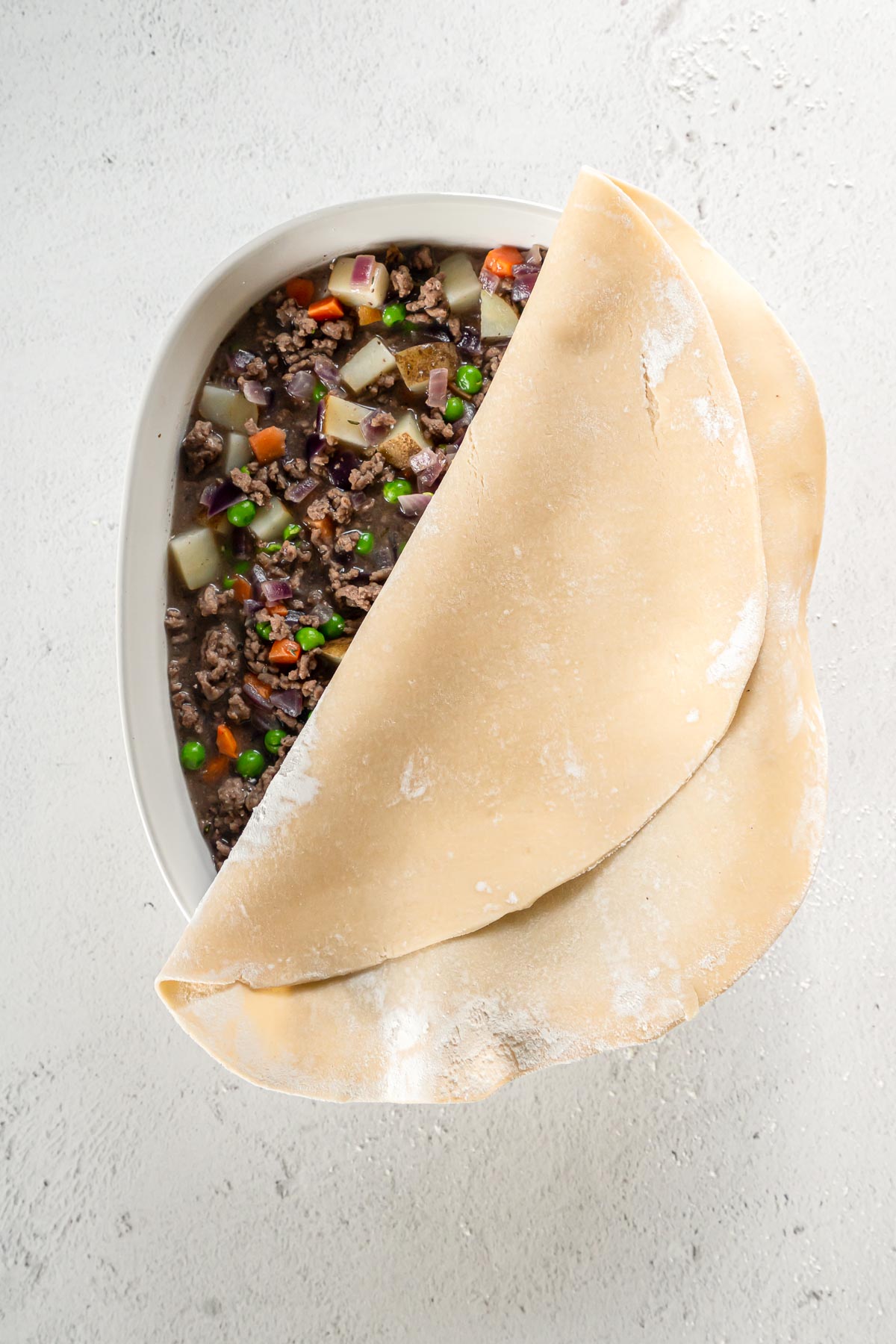 Pie crust being draped over a casserole dish with ground beef filling.