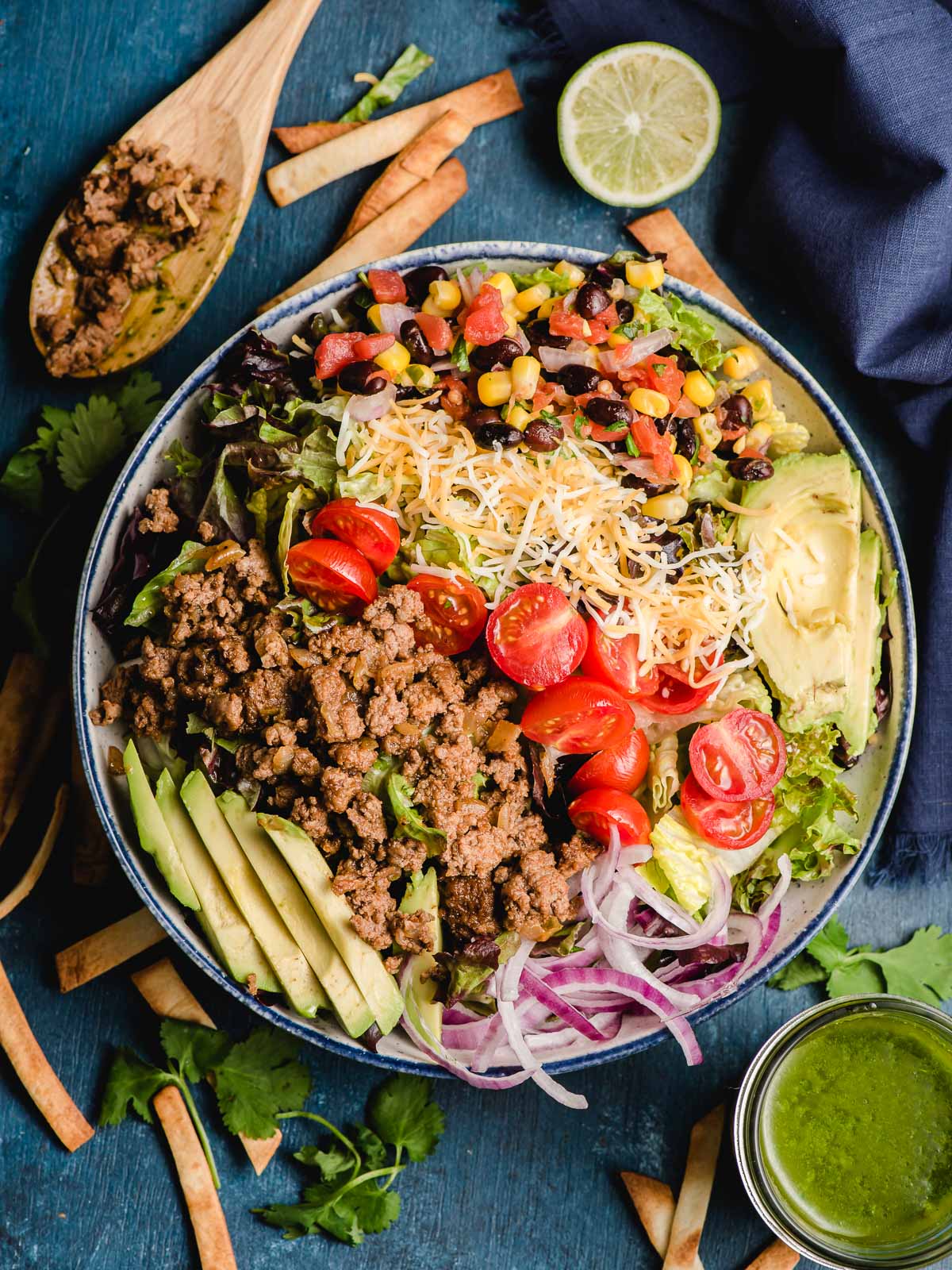 A colorful bowl of Ground Beef Taco Salad served on a table surrounded by tortilla strips, lime, and a spoon.