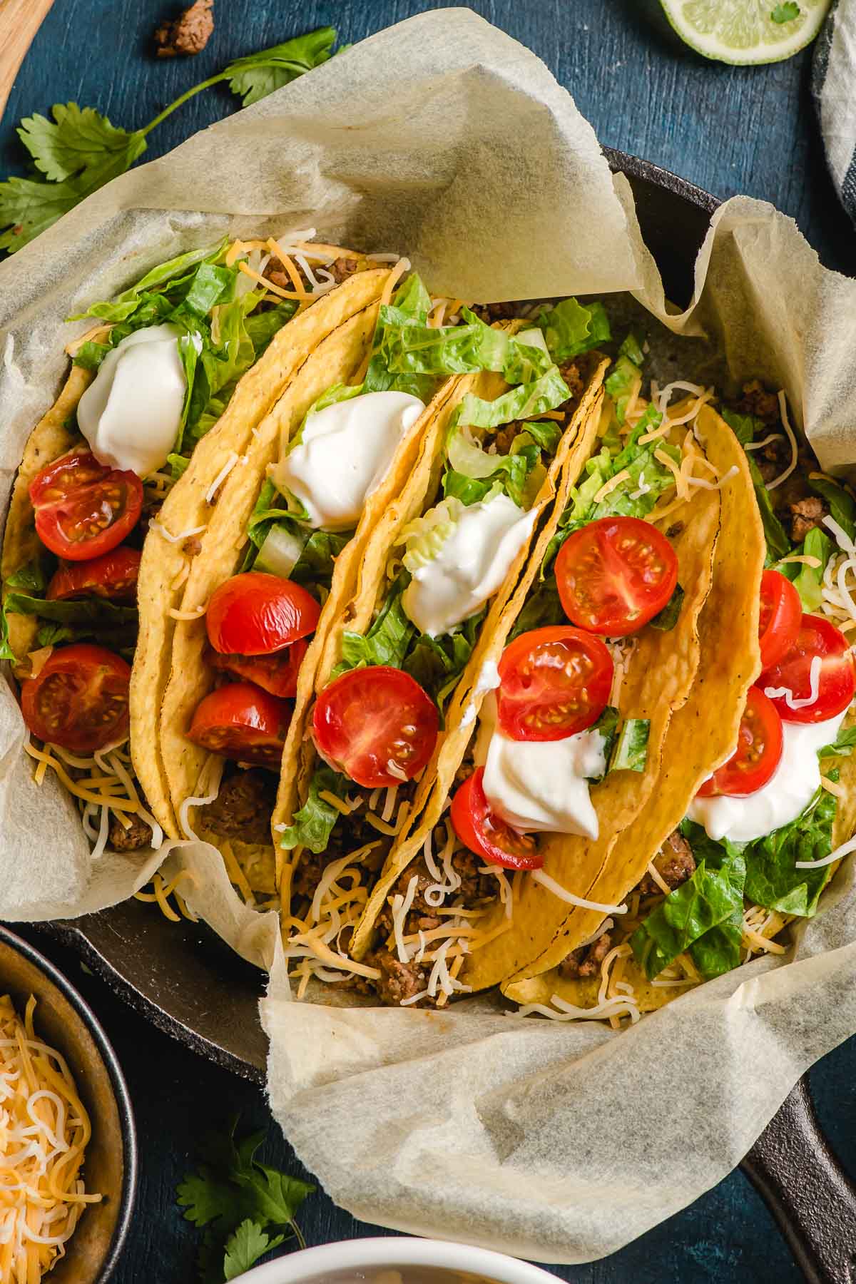 Five crunchy ground beef tacos in a skillet topped with tomatoes, shredded lettuce, and a dollop of sour cream.