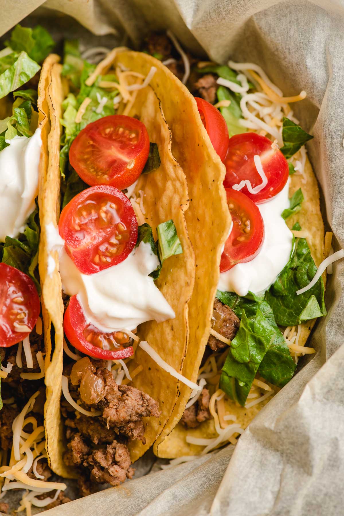 Three crunchy tacos topped with ground beef, tomatoes, sour cream, and lettuce in a brown paper lined skillet.