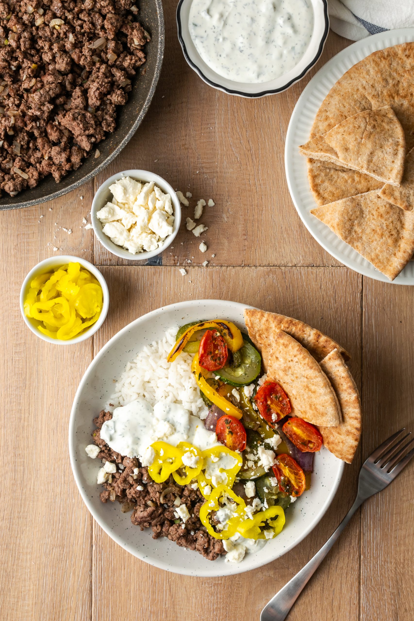 Ground Beef Gyro Bowls shown with bowls of banana peppers, feta, and tzatziki on the side.