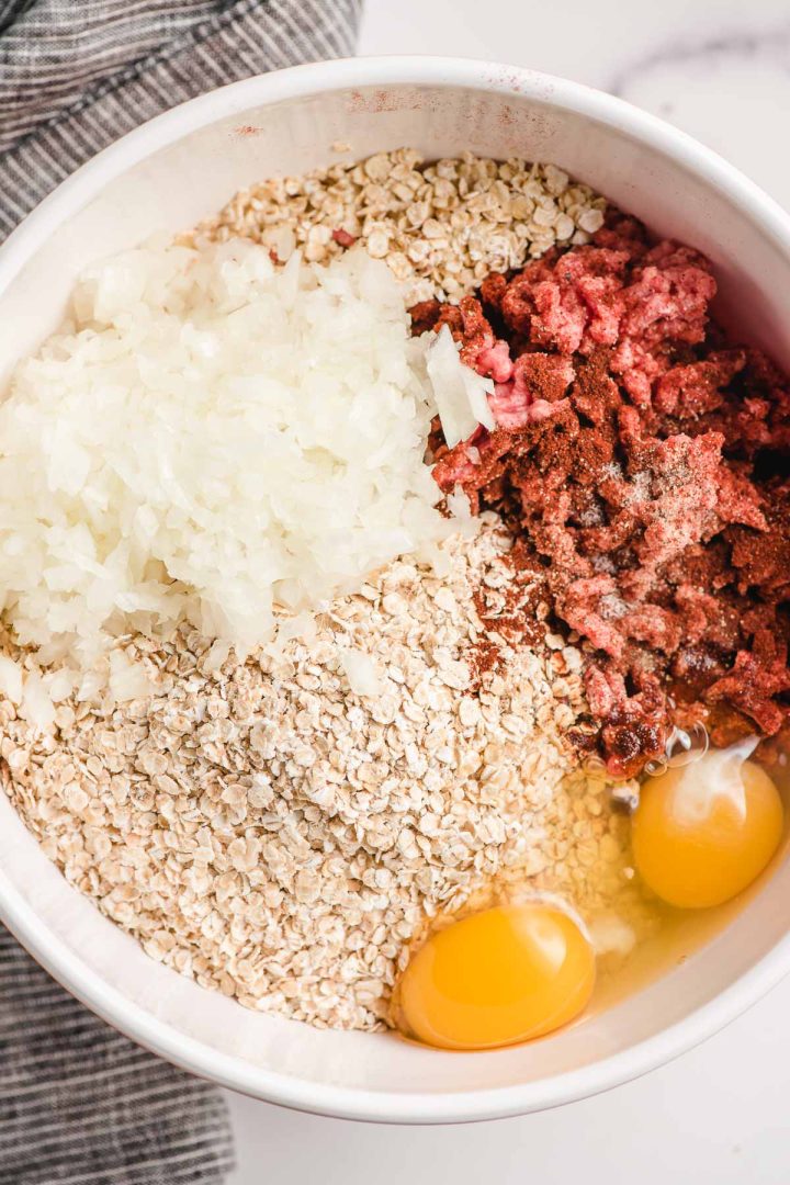 Bowl with ingredients for homemade meatballs--ground beef, quick oats, eggs, onion, evaporated milk, and spices.