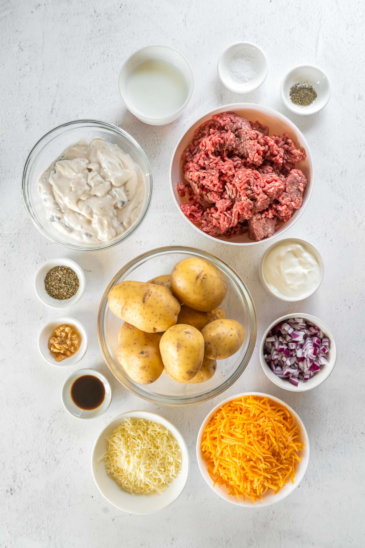 Ingredients displayed in bowls- yukon gold potatoes, ground beef, condensed soup, shredded cheese, garlic, milk, sour cream, worcestershire sauce, salt and pepper.