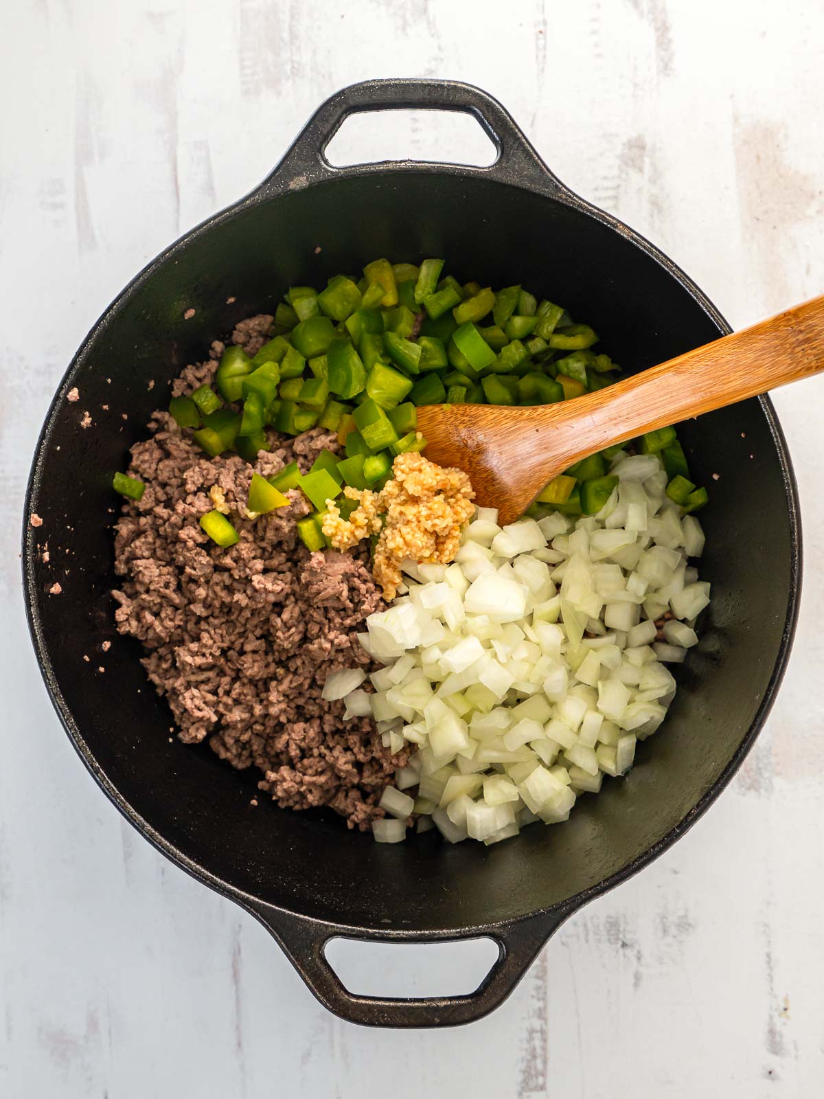 Wooden spatula stirring ground beef, onions, green pepper, and garlic in a cast iron pot.