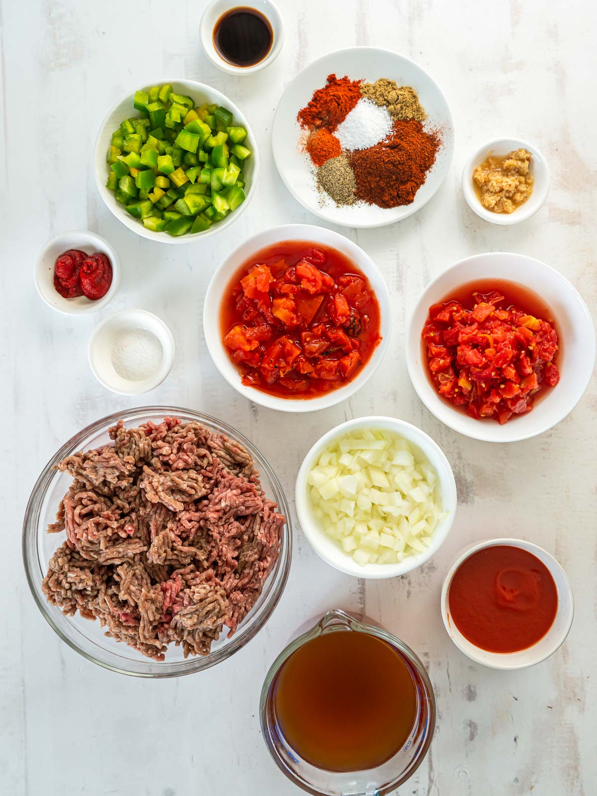 Ingredients for chili in bowls- ground beef, onions, green peppers, chopped tomatoes, spices, garlic, tomato paste, and broth.