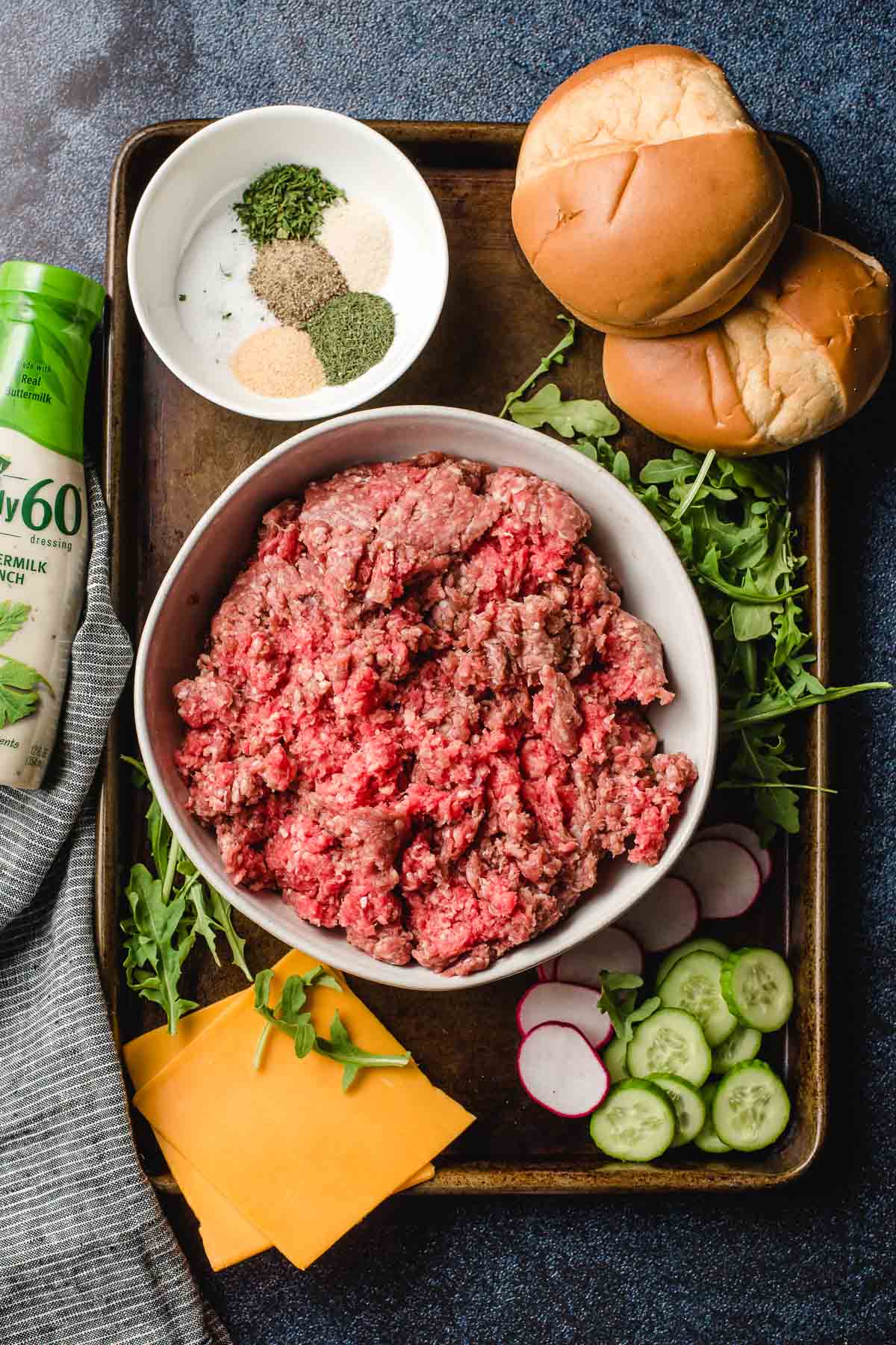 Sheet pan filled with ingredients to make burgers- ground beef, salt and pepper and herbs, cheese slices, arugula, buns, and sliced cucumber and radishes.