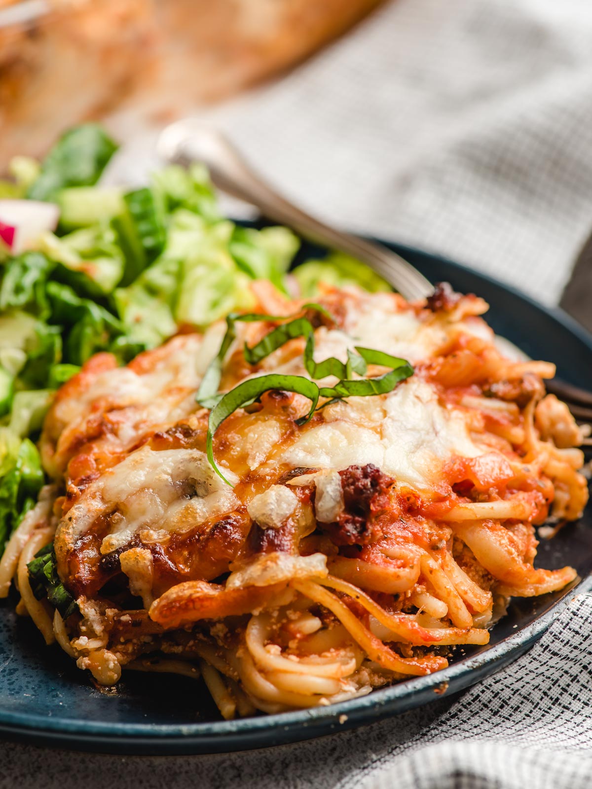 Baked spaghetti casserole served on a navy blue plate with salad greens.