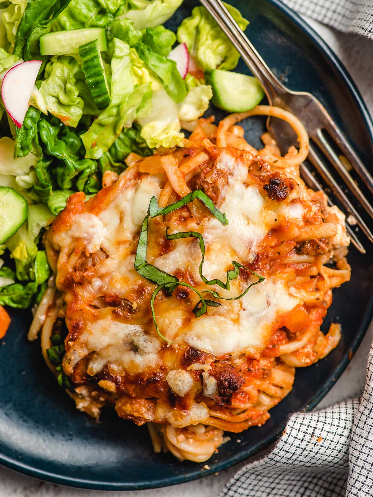 Cheesy baked spaghetti on a blue plate with salad greens on the side.