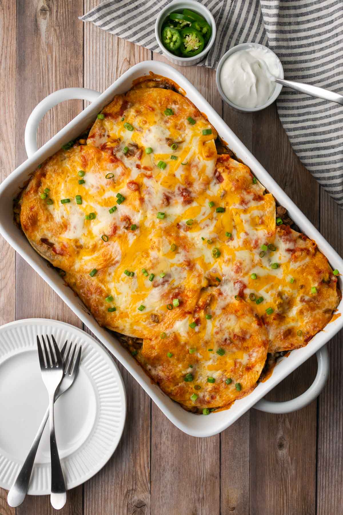Baked tortilla lasagna in a white casserole dish, with bowls of sour cream and jalapenos on the side.