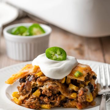 Taco lasagna shown on a white plate topped with sour cream and a slice of jalapeno.