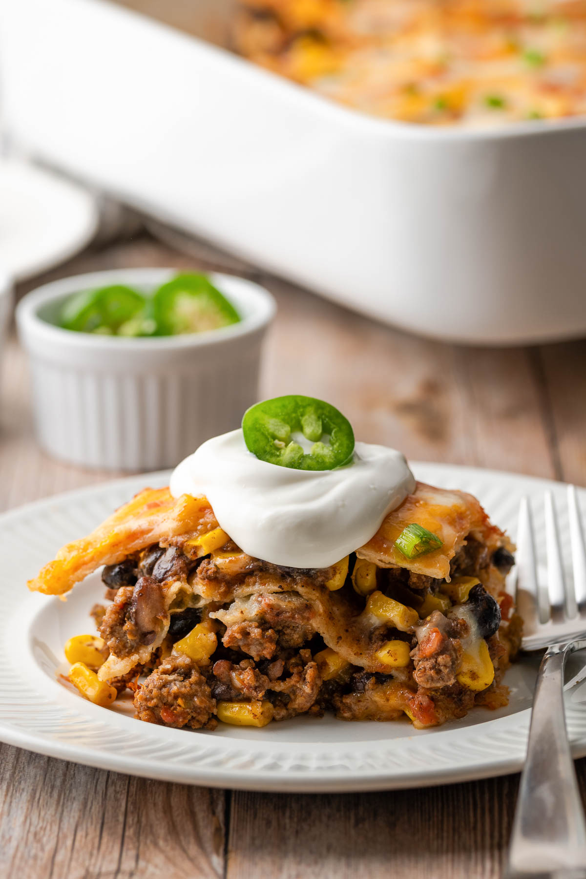 Taco lasagna shown on a white plate topped with sour cream and a slice of jalapeno.